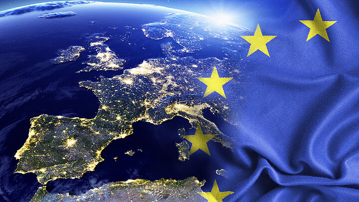 Grafika prezentująca fragment terytorium Europy widziane nocą z przestrzeni kosmicznej z ogniskami świateł w największych aglomeracjach oraz fragmentem flagi Unii Europejskiej – żółtych gwiazd ułożonych w okrąg na niebieskim tle.