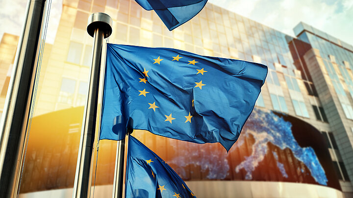 Zdjęcie flag Unii Europejskiej (żółte gwiazdy ułożone w okręgu na niebieskim tle) na tle nowoczesnego budynku o szklanej elewacji, w której odbija się słońce.